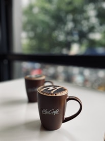 Two brown coffee mugs with a logo sit on a white surface. The mugs are topped with a frothy layer, likely featuring cocoa powder art. In the background, there is a blurred view of green foliage and a window, suggesting an indoor cafe setting.