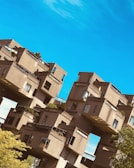 A complex arrangement of modular, cubic residences stacked in an artistic, geometric pattern against a clear blue sky. The structure features earthy tones with visible windows, balconies, and rooftop greenery.