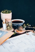 A variety of items are arranged on a wooden surface, including a blue and white patterned bowl filled with a dark liquid, a jar with a cork lid containing dried herbs, a small bottle with a dropper, and a tea infuser. There are also wooden coasters and a wooden spoon, along with some loose leaf greens and a document in the foreground.