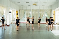 Several individuals dressed in black dance attire are practicing in a bright and spacious dance studio with mirrored walls and large windows adorned with sheer curtains. They appear to be engaged in various dance poses and movements, suggesting a dance class or rehearsal.