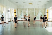 Several individuals dressed in black dance attire are practicing in a bright and spacious dance studio with mirrored walls and large windows adorned with sheer curtains. They appear to be engaged in various dance poses and movements, suggesting a dance class or rehearsal.