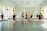 Several individuals dressed in black dance attire are practicing in a bright and spacious dance studio with mirrored walls and large windows adorned with sheer curtains. They appear to be engaged in various dance poses and movements, suggesting a dance class or rehearsal.
