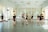 Several individuals dressed in black dance attire are practicing in a bright and spacious dance studio with mirrored walls and large windows adorned with sheer curtains. They appear to be engaged in various dance poses and movements, suggesting a dance class or rehearsal.