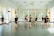 Several individuals dressed in black dance attire are practicing in a bright and spacious dance studio with mirrored walls and large windows adorned with sheer curtains. They appear to be engaged in various dance poses and movements, suggesting a dance class or rehearsal.