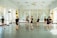 Several individuals dressed in black dance attire are practicing in a bright and spacious dance studio with mirrored walls and large windows adorned with sheer curtains. They appear to be engaged in various dance poses and movements, suggesting a dance class or rehearsal.