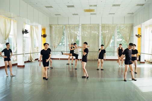 Several individuals dressed in black dance attire are practicing in a bright and spacious dance studio with mirrored walls and large windows adorned with sheer curtains. They appear to be engaged in various dance poses and movements, suggesting a dance class or rehearsal.