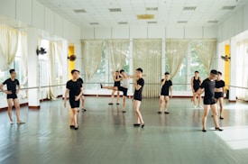 Several individuals dressed in black dance attire are practicing in a bright and spacious dance studio with mirrored walls and large windows adorned with sheer curtains. They appear to be engaged in various dance poses and movements, suggesting a dance class or rehearsal.
