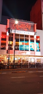 A brightly illuminated urban storefront featuring neon red and white signage for 'Kentucky,' a pizza restaurant established in 1942 in Buenos Aires. The facade includes large glass windows displaying the interior where people are dining. Street lights and traffic signals are visible in front of the building, enhancing the lively night-time atmosphere.