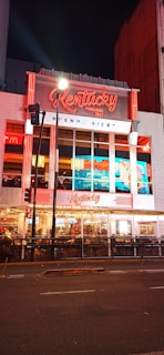 A brightly illuminated urban storefront featuring neon red and white signage for 'Kentucky,' a pizza restaurant established in 1942 in Buenos Aires. The facade includes large glass windows displaying the interior where people are dining. Street lights and traffic signals are visible in front of the building, enhancing the lively night-time atmosphere.