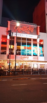 A brightly illuminated urban storefront featuring neon red and white signage for 'Kentucky,' a pizza restaurant established in 1942 in Buenos Aires. The facade includes large glass windows displaying the interior where people are dining. Street lights and traffic signals are visible in front of the building, enhancing the lively night-time atmosphere.
