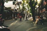 A bustling urban street lined with trees and local businesses is busy with activity. Motorbikes, some carrying cargo, navigate the road, while people engage with the environment. The sunlight filters through the trees, creating a lively atmosphere.