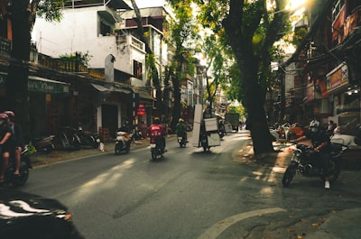 A bustling urban street lined with trees and local businesses is busy with activity. Motorbikes, some carrying cargo, navigate the road, while people engage with the environment. The sunlight filters through the trees, creating a lively atmosphere.