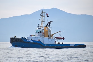 A tugboat named 'HORCON' is seen gliding through calm waters. The boat has a blue hull with a white and yellow cabin structure. Flags are mounted atop the mast, and a small lifeboat is secured on deck. In the background, a large, misty mountain forms the backdrop under an overcast sky.