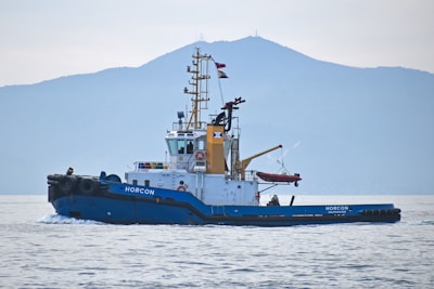 A tugboat named 'HORCON' is seen gliding through calm waters. The boat has a blue hull with a white and yellow cabin structure. Flags are mounted atop the mast, and a small lifeboat is secured on deck. In the background, a large, misty mountain forms the backdrop under an overcast sky.