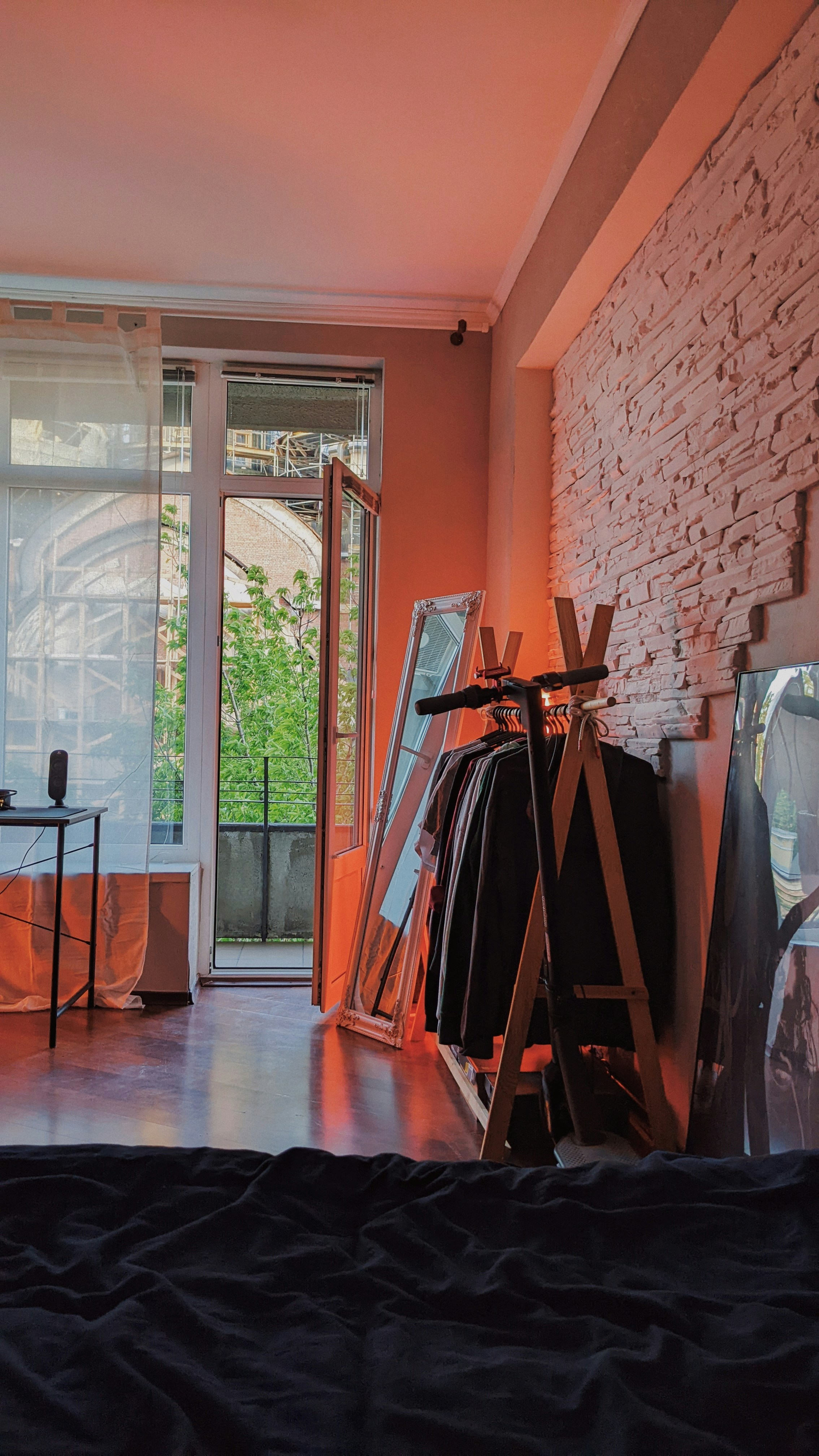 A cozy room features a bed in the foreground, with a stylishly arranged clothes rack and large mirror against a textured white brick wall. Soft natural light filters through an open glass door leading to a balcony, revealing lush greenery outside. A minimalist desk with a small lamp is situated near the window.