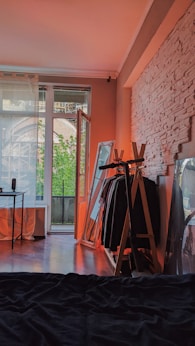 A cozy room features a bed in the foreground, with a stylishly arranged clothes rack and large mirror against a textured white brick wall. Soft natural light filters through an open glass door leading to a balcony, revealing lush greenery outside. A minimalist desk with a small lamp is situated near the window.