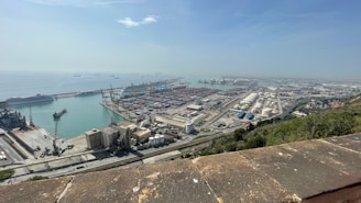 A large industrial port area with numerous shipping containers, cranes, and several docked ships along a vast waterfront. The area includes storage tanks and various industrial buildings. The ocean extends to the horizon with distant ships visible on the water. The foreground has a stone or concrete ledge, and the landscape includes some greenery.