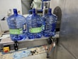 Three large blue plastic water jugs are placed on a conveyor belt inside a bottling or filtration facility. The jugs have labels with green and white text. The surrounding area features industrial equipment, with some pipes and a large container visible in the background. The floor has spilled water or possibly fragments of plastic.