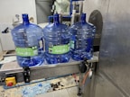 Three large blue plastic water jugs are placed on a conveyor belt inside a bottling or filtration facility. The jugs have labels with green and white text. The surrounding area features industrial equipment, with some pipes and a large container visible in the background. The floor has spilled water or possibly fragments of plastic.