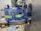 Three large blue plastic water jugs are placed on a conveyor belt inside a bottling or filtration facility. The jugs have labels with green and white text. The surrounding area features industrial equipment, with some pipes and a large container visible in the background. The floor has spilled water or possibly fragments of plastic.