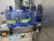 Three large blue plastic water jugs are placed on a conveyor belt inside a bottling or filtration facility. The jugs have labels with green and white text. The surrounding area features industrial equipment, with some pipes and a large container visible in the background. The floor has spilled water or possibly fragments of plastic.