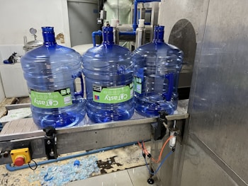 Three large blue plastic water jugs are placed on a conveyor belt inside a bottling or filtration facility. The jugs have labels with green and white text. The surrounding area features industrial equipment, with some pipes and a large container visible in the background. The floor has spilled water or possibly fragments of plastic.