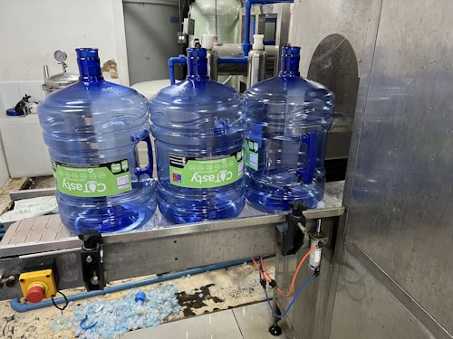 Three large blue plastic water jugs are placed on a conveyor belt inside a bottling or filtration facility. The jugs have labels with green and white text. The surrounding area features industrial equipment, with some pipes and a large container visible in the background. The floor has spilled water or possibly fragments of plastic.