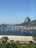 Several sailboats are anchored in a calm bay with a sandy beach in the foreground. The iconic Sugarloaf Mountain rises prominently in the background under a clear blue sky. Lush green trees border the beach alongside a roadway.