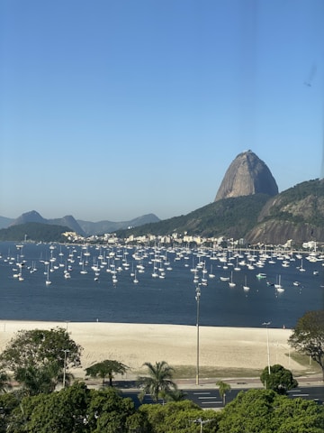 Several sailboats are anchored in a calm bay with a sandy beach in the foreground. The iconic Sugarloaf Mountain rises prominently in the background under a clear blue sky. Lush green trees border the beach alongside a roadway.