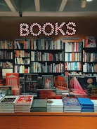 A bookstore interior features a glowing 'BOOKS' sign with circular lights. Below, shelves are filled with various genres, including graphic design, music, and interior design. The display table in front showcases an assortment of colorful books and magazines, including works on art, fashion, and architecture.