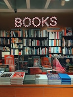 A bookstore interior features a glowing 'BOOKS' sign with circular lights. Below, shelves are filled with various genres, including graphic design, music, and interior design. The display table in front showcases an assortment of colorful books and magazines, including works on art, fashion, and architecture.