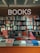 A bookstore interior features a glowing 'BOOKS' sign with circular lights. Below, shelves are filled with various genres, including graphic design, music, and interior design. The display table in front showcases an assortment of colorful books and magazines, including works on art, fashion, and architecture.