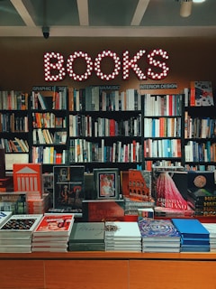 A bookstore interior features a glowing 'BOOKS' sign with circular lights. Below, shelves are filled with various genres, including graphic design, music, and interior design. The display table in front showcases an assortment of colorful books and magazines, including works on art, fashion, and architecture.