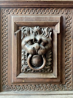 An intricately carved wooden lion head with a ring in its mouth, surrounded by ornate patterns in a rectangular frame. The craftsmanship showcases detailed fur and expressive features, highlighting the artistic skill involved in creating this decorative piece.