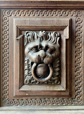 An intricately carved wooden lion head with a ring in its mouth, surrounded by ornate patterns in a rectangular frame. The craftsmanship showcases detailed fur and expressive features, highlighting the artistic skill involved in creating this decorative piece.