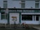 A storefront of a pork butcher shop with the name 'F. Heterich's Pork Butchers' prominently displayed. The facade includes an image of a pig with the words 'Pork Butcher' beneath it. Two bicycles are parked in front of the shop. A 'No Parking' sign is present, allowing loading for 30 minutes. The shop windows display additional text indicating sausage manufacturing.