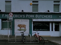 A storefront of a pork butcher shop with the name 'F. Heterich's Pork Butchers' prominently displayed. The facade includes an image of a pig with the words 'Pork Butcher' beneath it. Two bicycles are parked in front of the shop. A 'No Parking' sign is present, allowing loading for 30 minutes. The shop windows display additional text indicating sausage manufacturing.