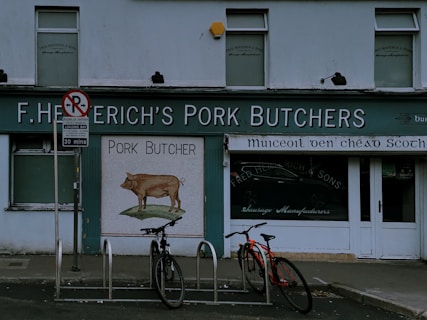 A storefront of a pork butcher shop with the name 'F. Heterich's Pork Butchers' prominently displayed. The facade includes an image of a pig with the words 'Pork Butcher' beneath it. Two bicycles are parked in front of the shop. A 'No Parking' sign is present, allowing loading for 30 minutes. The shop windows display additional text indicating sausage manufacturing.