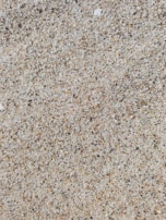 A close-up view of sand composed of tiny grains with a variety of colors including beige, brown, and white, creating a textured and granular appearance.
