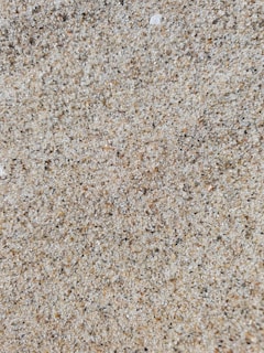 A close-up view of sand composed of tiny grains with a variety of colors including beige, brown, and white, creating a textured and granular appearance.