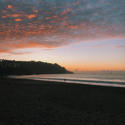 The image captures a serene beach at sunset with a vast expanse of sand in the foreground and gentle waves lapping at the shore. The coastline is silhouetted against a vibrant sky, which transitions from deep blues and purples to warm oranges and pinks. Scattered clouds add texture to the sky, and a few distant figures are visible along the shoreline.