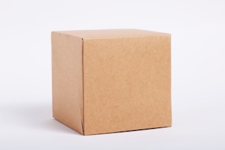 A plain, brown cardboard box placed against a light gray background. The box appears simple and unadorned, showing sharp edges and a smooth surface.