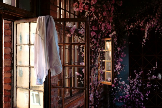 A white shirt hangs on the corner of a rustic wooden window lit by soft natural light. The surrounding walls are made of brick, giving a vintage feel. In the background, a plethora of pink blossoms adorn the space, adding a touch of vibrant color. The scene is tranquil and intimate, evoking a poetic atmosphere with the interplay of shadows and warm sunlight.