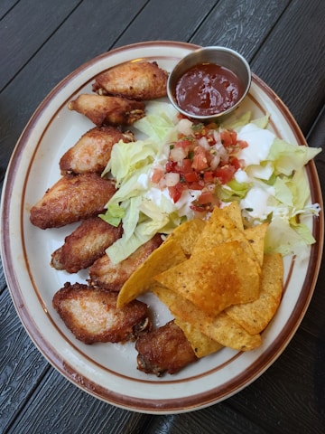 A plate of food featuring crispy chicken wings, a serving of tortilla chips, lettuce, diced tomatoes, sour cream, and a small dish of red dipping sauce.