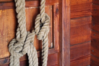 A thick, textured rope is tied in a complex knot against a wooden wall. The wall features rich, dark brown wooden planks with visible grains and knots, suggesting a rustic or nautical setting.