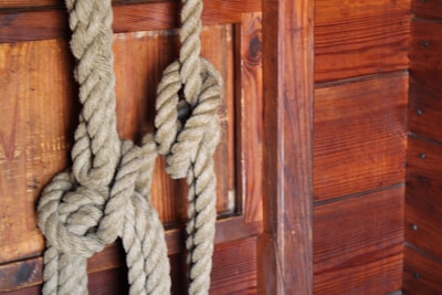 A thick, textured rope is tied in a complex knot against a wooden wall. The wall features rich, dark brown wooden planks with visible grains and knots, suggesting a rustic or nautical setting.