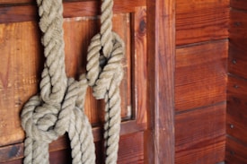 A thick, textured rope is tied in a complex knot against a wooden wall. The wall features rich, dark brown wooden planks with visible grains and knots, suggesting a rustic or nautical setting.