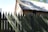 A rustic, thatched-roof house sits behind a tall, pointed wooden fence. The wooden structure displays an earthy, historical character with shadows cast across both the roof and the fence. Some greenery is visible in the foreground, adding a touch of nature to the scene.