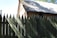 A rustic, thatched-roof house sits behind a tall, pointed wooden fence. The wooden structure displays an earthy, historical character with shadows cast across both the roof and the fence. Some greenery is visible in the foreground, adding a touch of nature to the scene.