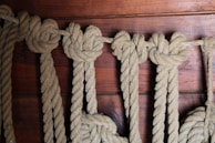 Several thick, tan ropes are neatly knotted and hanging against a wooden background with a rich, reddish-brown hue. The knots are evenly spaced and the rough texture of the ropes is clearly visible, creating a rustic and nautical feel.